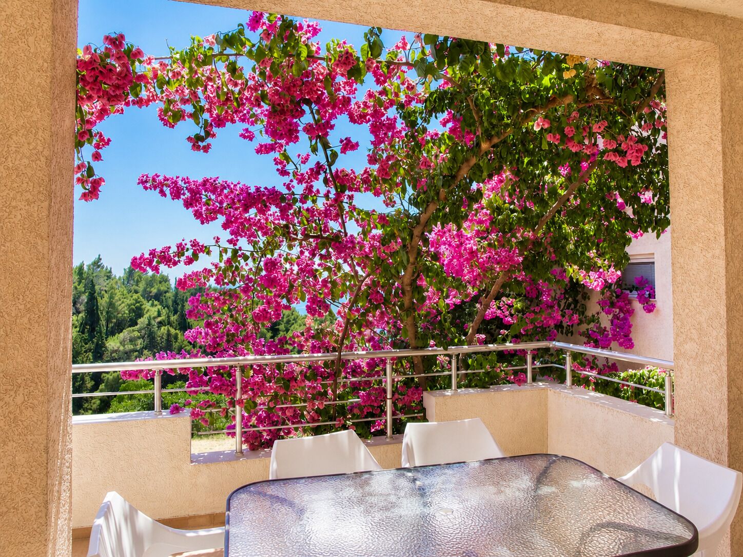 A balcony with a glass table and white chairs, overlooking a tree with pink flowers.