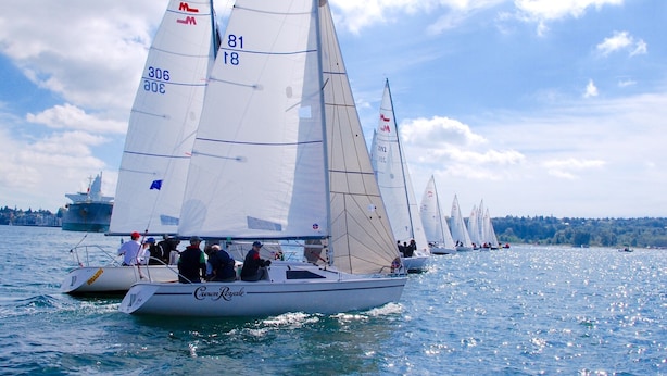 Un velero con varias velas, una tripulación y un barco grande al fondo.