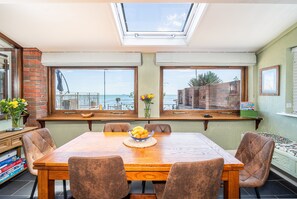 Un comedor con una mesa de madera, cuatro sillas, un jarrón de flores y vista al mar a través de amplios ventanales.