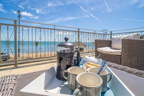 Un balcón con vista al mar, un sofá de mimbre, una mesa con una cafetera, tazas y pan.