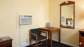 A hotel room with a wooden chair, a small table, a mirror, and a wall-mounted air conditioner.
