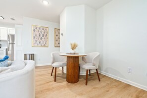 Een moderne woonkamer met een ronde houten tafel, twee witte stoelen, een bank en een keuken in de achtergrond.