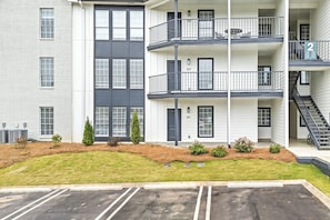 A multi-story residential building with balconies, a parking lot, and a small landscaped area with shrubs. - First Floor 3 BR Condo ~ Newly Renovated, Sleeps 8 (Auburn)