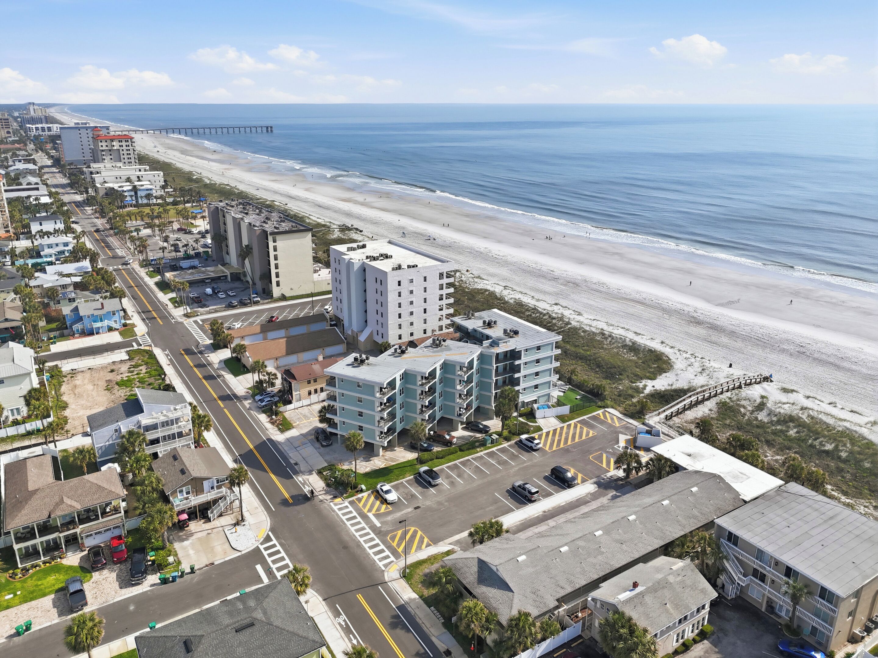 An aerial view of a coastal area with a beach, a pier, and a multi-story building with balconies.