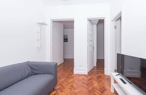 Ein modernes Wohnzimmer mit einem grauen Sofa, einem Flachbildfernseher auf einem weißen Schrank und einem Holzboden mit Fischgrätmuster.