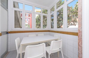 Ein Eckzimmer mit weißem Tisch und Stühlen, großen Fenstern und Blick auf Gebäude und Bäume.