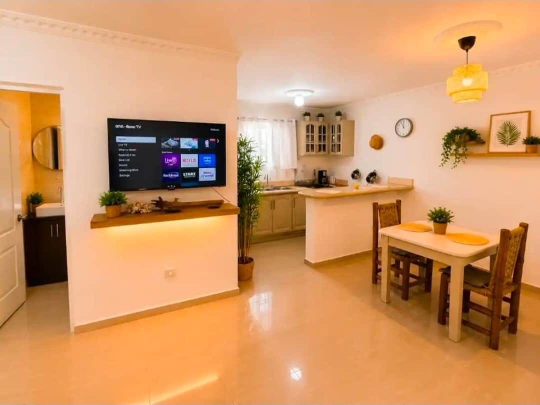 A modern living room with a flat-screen TV, a dining area with wooden chairs, and a kitchen in the background.