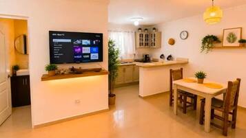A modern living room with a flat-screen TV, a dining area with wooden chairs, and a kitchen in the background.