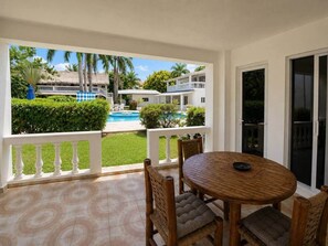 Un espacio al aire libre cubierto con una mesa y sillas de madera redonda, una barandilla blanca, y vistas a una piscina y edificios al fondo.