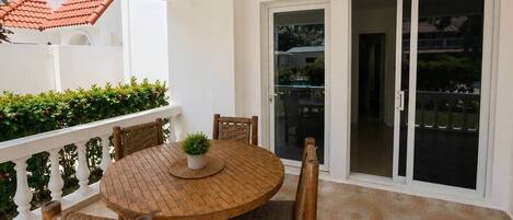 A balcony with a round wooden table and wicker chairs, a potted plant, and a sliding glass door leading to a covered area.