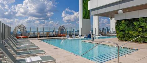A rooftop pool with lounge chairs, a glass balustrade, and a city view.