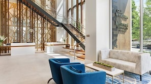 Un moderno vestíbulo de hotel con mostrador de recepción, sillas azules, un sofá, una escalera y ventanas amplias.