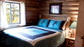 A log cabin bedroom with a bed, a window with curtains, a framed picture on the wall, and a nightstand with a lamp. - Cowboy Log Cabin, Mountain views and Firepit (Bigfork)