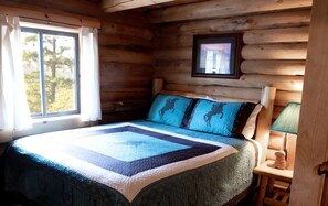 Ein Schlafzimmer in einer Blockhütte mit einem Bett, einem Fenster mit Vorhängen, einem gerahmten Bild an der Wand und einem Nachttisch mit einer Lampe.