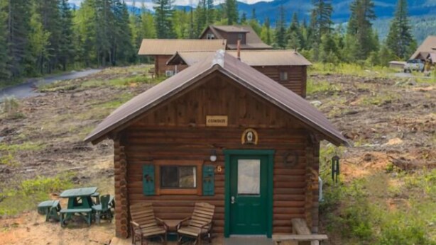 A rustic log cabin with a green door, a small porch with wooden chairs, and a sign above the door. - Cowboy Log Cabin, Mountain views and Firepit (Bigfork)