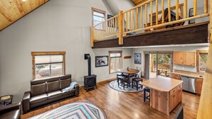 A spacious loft with a high ceiling, a wooden staircase, a central island with seating, a built-in stove, and a kitchen area. - Experience Montana Cabins - 9 homes on 15 acres (Bigfork)