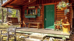 A rustic log cabin with a green door, a bench, a chair, and a hanging flower basket. - Experience Montana Cabins - 9 homes on 15 acres (Bigfork)