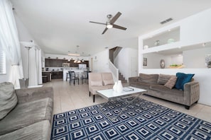A spacious living area with a sofa, coffee table, and patterned rug. A kitchen is visible in the background, and there is a staircase leading to another level.