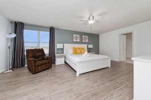A bedroom with a bed, a brown chair, a nightstand, a ceiling fan, and a view through a window.