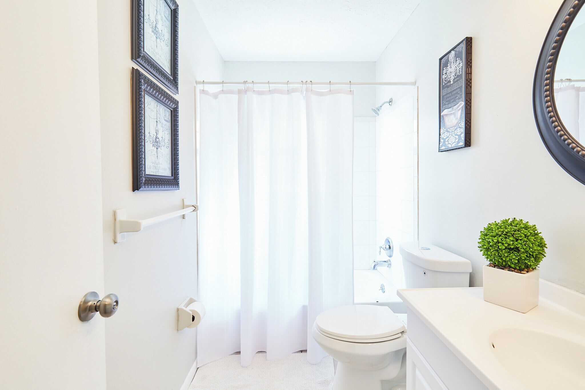 Ein Badezimmer mit Toilette, Waschbecken und Dusche. An den Wänden hängen Bilder in Rahmen und auf dem Waschbecken steht ein kleiner Topfpflanze.