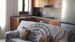 A living room with a sofa, a kitchen area with wooden cabinets, a refrigerator, and a window with curtains. - El Mirador Alto Apartment (Trevélez)