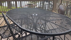 A round metal table with chairs on an outdoor deck with a view of a lake and trees.