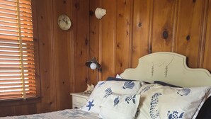 A wooden paneled room with a bed, a nightstand, a lamp, and a window with blinds.