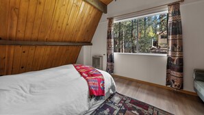 Un dormitorio con techo de madera, una cama con una manta colorida, una ventana con cortinas y vistas a árboles.