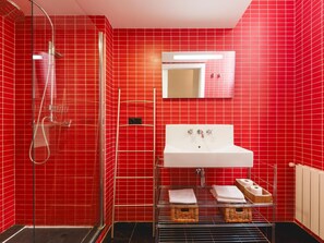 Une salle de bain avec des murs recouverts de carreaux rouges, une douche en verre, un lavabo blanc avec un miroir au-dessus, et un porte-serviettes avec des serviettes pliées et des paniers.