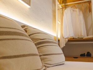 Un lit avec des oreillers à rayures, une table de chevet et une armoire suspendue contenant des vêtements et des chaussures.