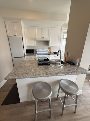 A kitchen with white cabinets, a granite countertop, a built-in oven, a microwave, and two bar stools.