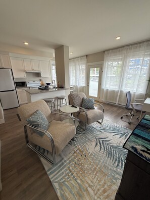 A modern kitchen with white cabinets, a stove, and a refrigerator. There is a dining area with a table and chairs, and a living room with a sofa and a coffee table.