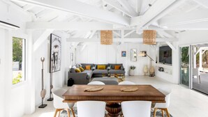 Ruime kamer met een houten eettafel, witte stoelen, een grijze bank met kleurrijke kussens en een groot raam.