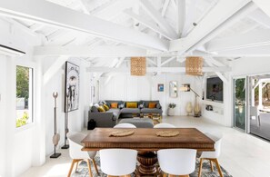 Ein geräumiges Zimmer mit einem Holzespeisetisch, weißen Stühlen, einem grauen Sofa mit bunten Kissen und einem großen Fenster.