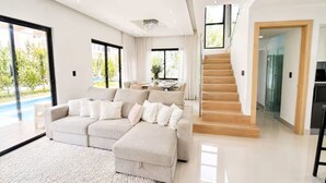 Un salon moderne avec un grand canapé d’angle beige, un escalier et une porte vitrée donnant sur un espace repas.