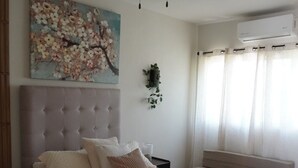 A bedroom with a bed, a ceiling fan, a wall-mounted air conditioner, a window with curtains, and a floral painting. - Central Zone Department (La Paz)