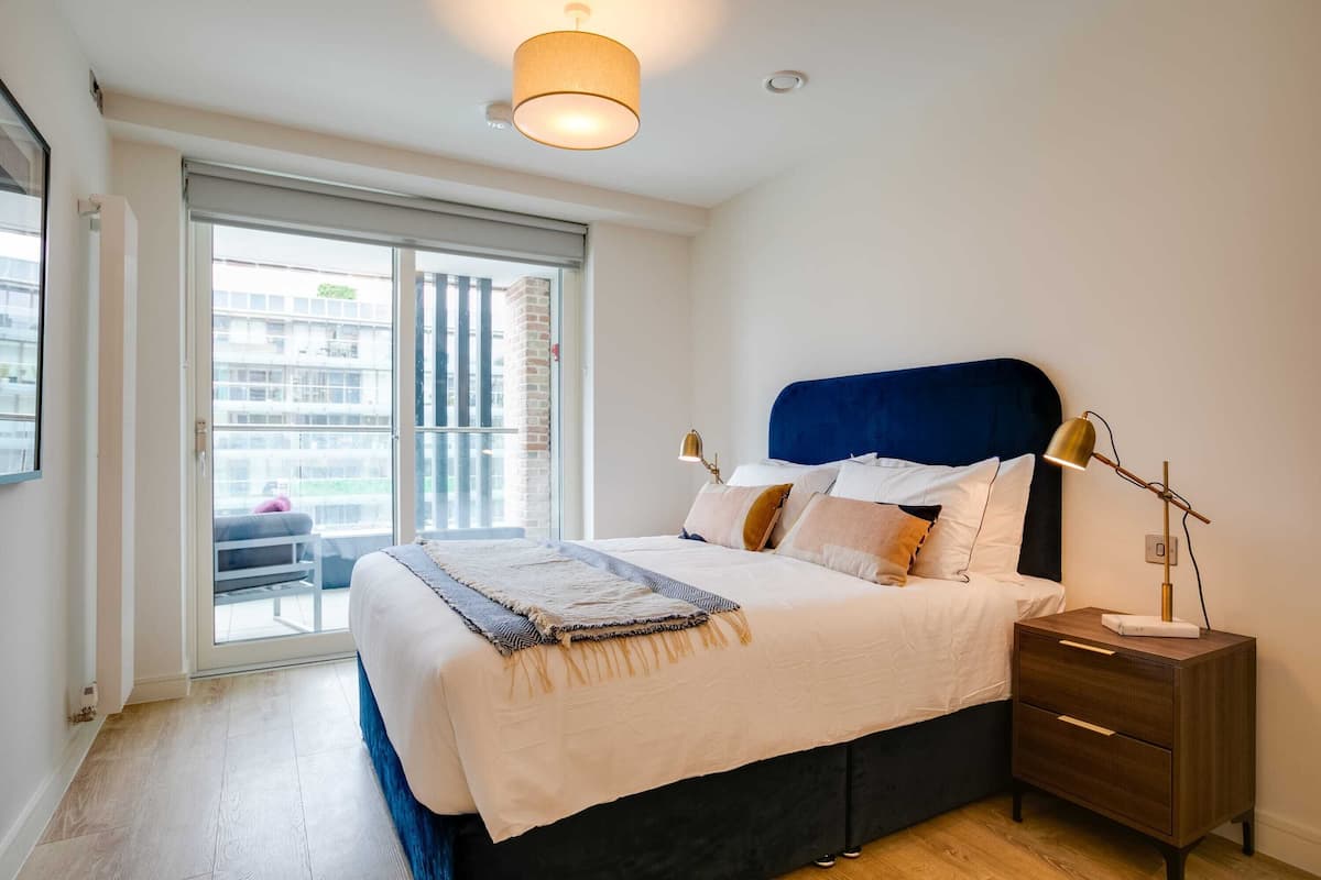 A bedroom with a bed, bedside table, lamp, and a view through a sliding door to an outdoor balcony.