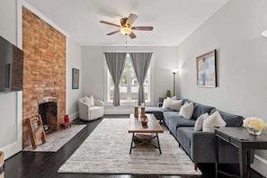 A living room with a brick wall, a fireplace, a ceiling fan, a sofa, a coffee table, and a view through a window. - Modern 3BR Home - FirePit + Grill - Sleeps 9 (Columbus)