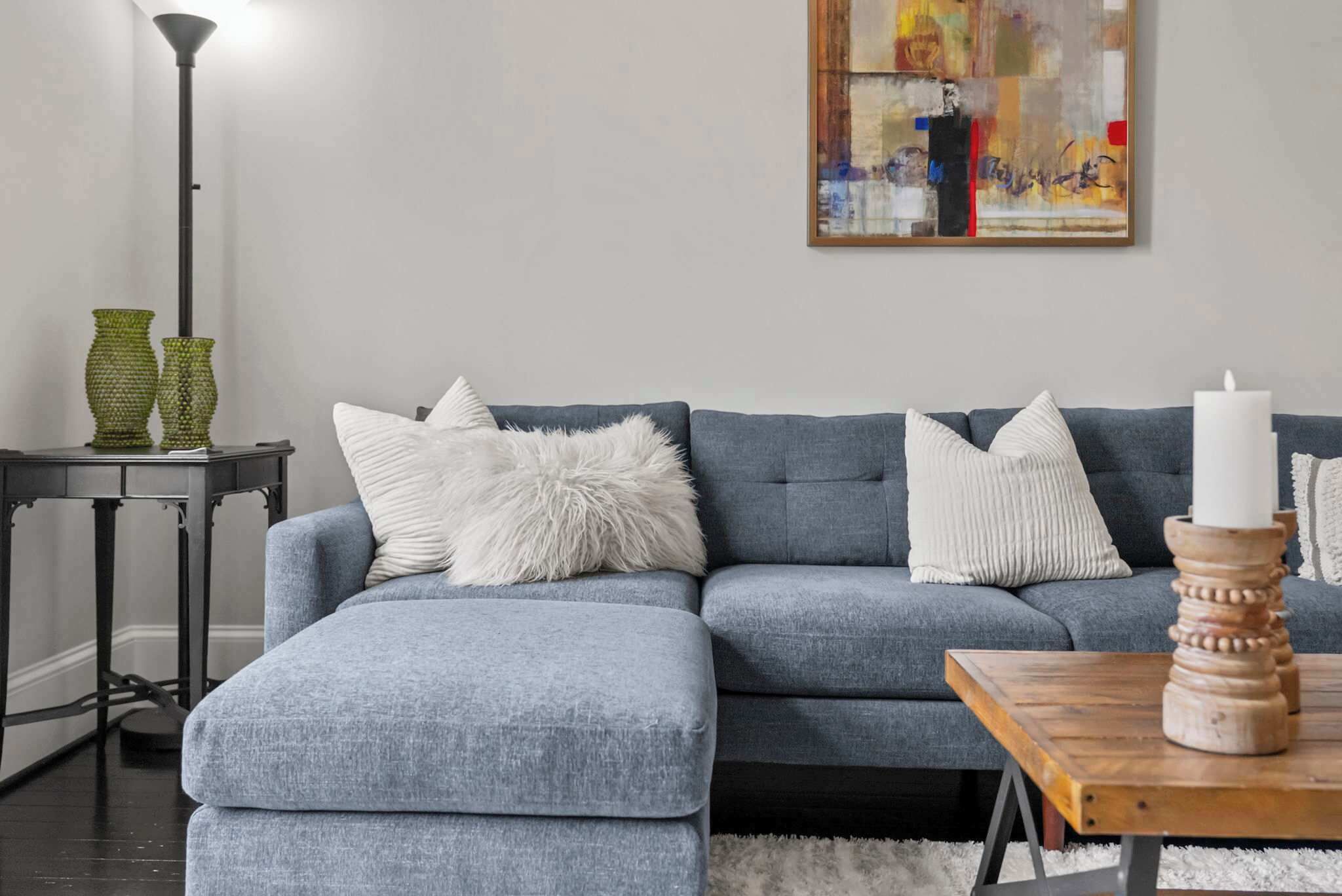 A living room with a grey sectional sofa, white fluffy pillows, a small side table with green vases, and a painting on the wall.
