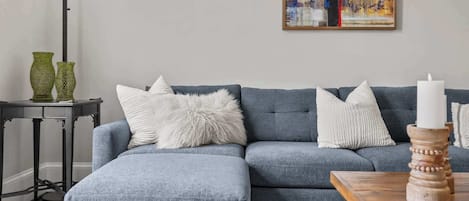 A living room with a grey sectional sofa, white fluffy pillows, a small side table with green vases, and a painting on the wall.