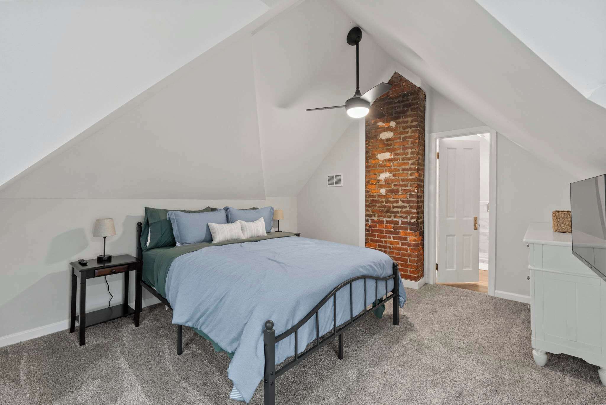 Une chambre avec un plafond en pente, un lit avec une tête de lit, une table de chevet, une lampe, une commode et un mur d’accent en briques.