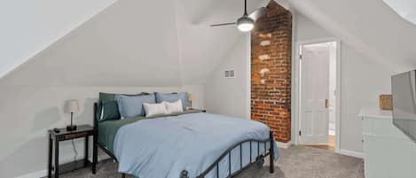 Une chambre avec un plafond en pente, un lit avec une tête de lit, une table de chevet, une lampe, une commode et un mur d’accent en briques.
