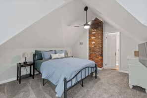 Una camera da letto con soffitto spiovente, un letto con testiera, un comodino, una lampada, una cassettiera e una parete decorativa in mattoni.