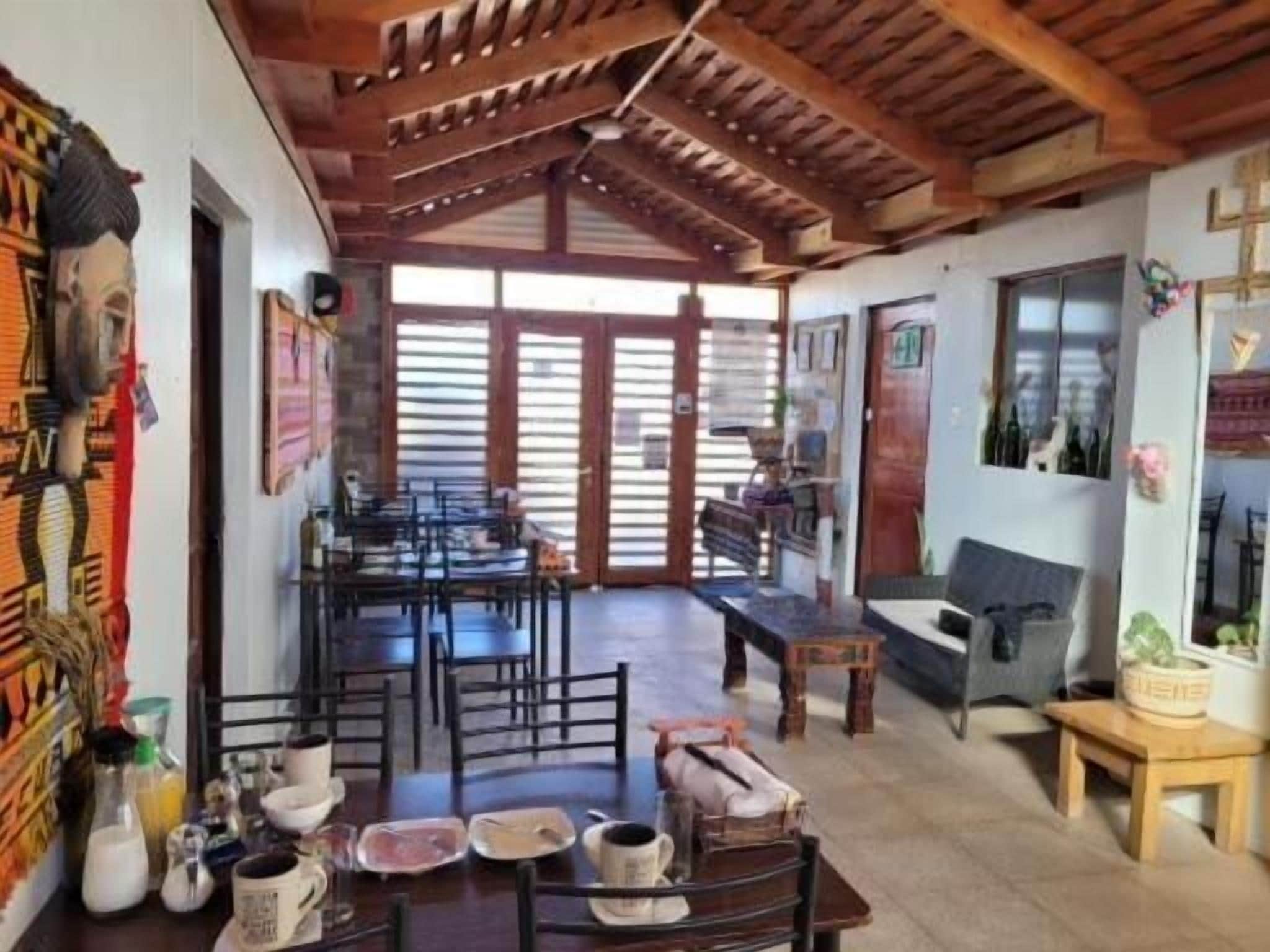 Un comedor con mesas y sillas de madera, una zona de cocina con refrigerador y alacenas, y un patio con techo de madera y ventanas amplias.