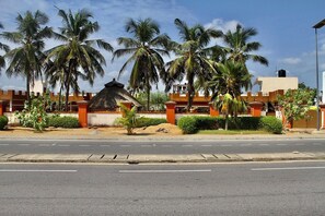 A street with palm trees, a building with a thatched roof, and a water tank on top. - Private Beach House Fidjrosse, 2 Steps to the Ocean (Cotonou)