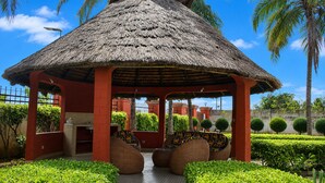 Een paviljoen met rieten dak en betegelde vloer, omgeven door verzorgde heggen en palmbomen.