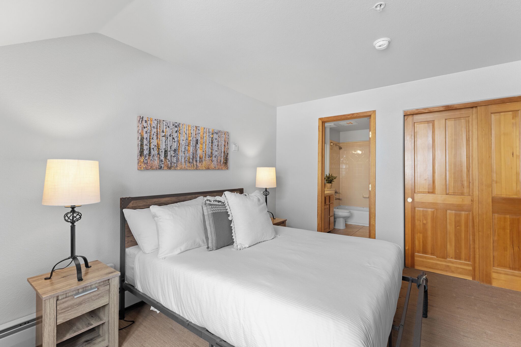 A bedroom with a bed, two bedside lamps, a wooden nightstand, a wooden wardrobe, and a bathroom visible through an open door.