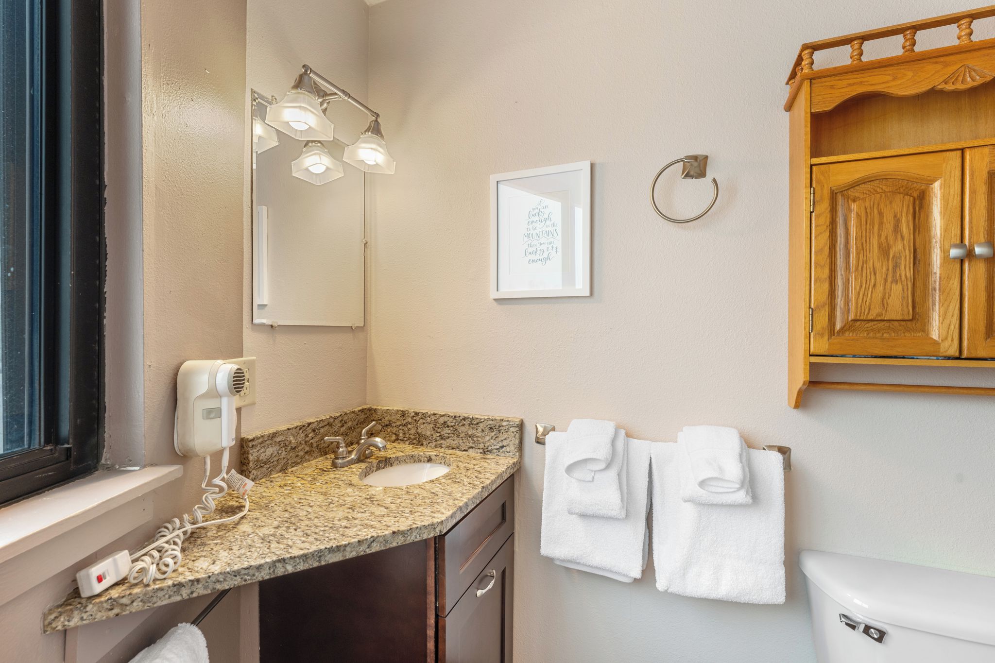 Une salle de bain avec un lavabo doté d’un plan de travail en granit, un sèche-cheveux, un miroir, une citation encadrée, un porte-serviettes avec des serviettes et des armoires en bois.