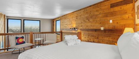 A hotel room with a large bed, a wooden wall, a balcony with a chair, and a view of mountains.