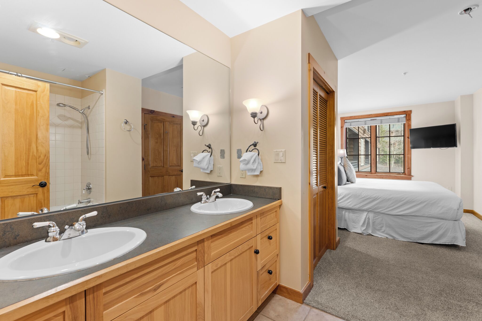 A bathroom with two sinks, a shower, a large mirror, and a bed in the adjacent room.